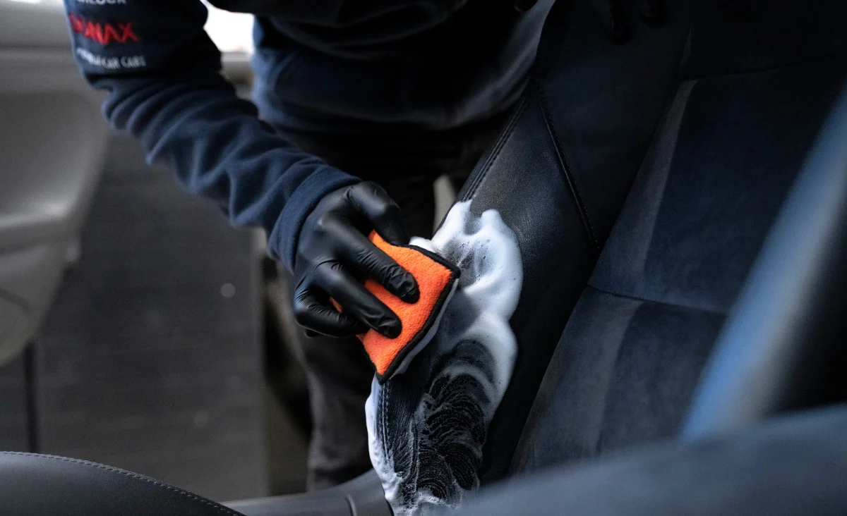 Hand mit schwarzem Handschuh reinigt mit oranger Schwammapplikator und weißem Schaum die schwarze Autotürinnenseite