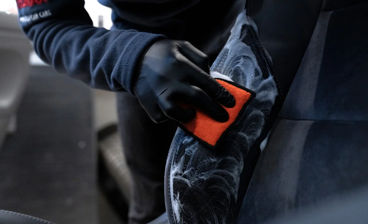 Hand mit schwarzem Handschuh reinigt mit oranger Schwammapplikator und weißem Schaum die schwarze Autotürinnenseite