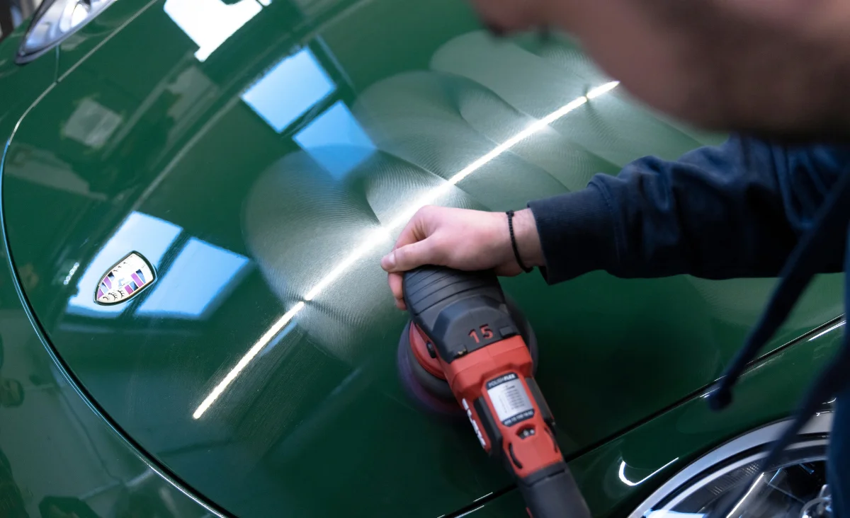 Nahaufnahme einer Hand, die mit einem Poliergerät die Motorhaube eines grünen Fahrzeugs mit Porsche-Emblem bearbeitet