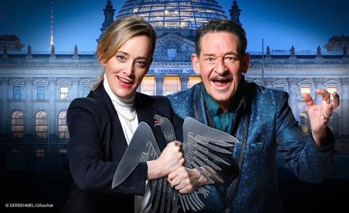 Zwei Personen mit rußverschmierten Gesichtern und erschrockenen Ausdrücken vor einem brennenden historischen Gebäude im Hintergrund