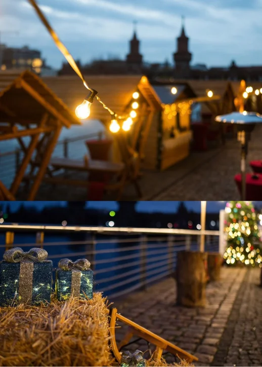 Abendlicher Blick über den All inclusive Weihnachtsmarkt an der Spree in Berlin: Beleuchtete Holzhütten und Lichterketten am Spreespeicher im Vordergrund, während im Hintergrund die Oberbaumbrücke mit einer fahrenden U-Bahn zu sehen ist.