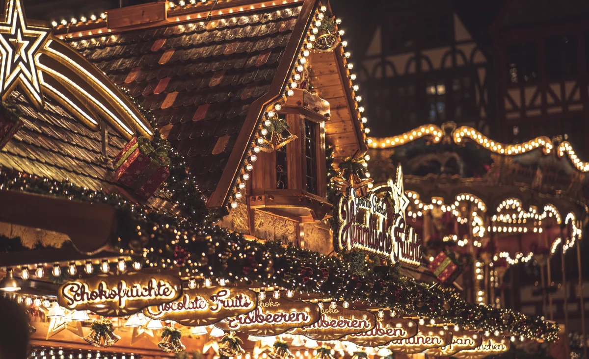 Nahaufnahme eines festlich beleuchteten Holzstandes auf dem Berliner Weihnachtszauber mit dekorativen Lichterketten und handgeschriebenen Schildern für Schokofrüchte, Erdbeeren und Beeren.