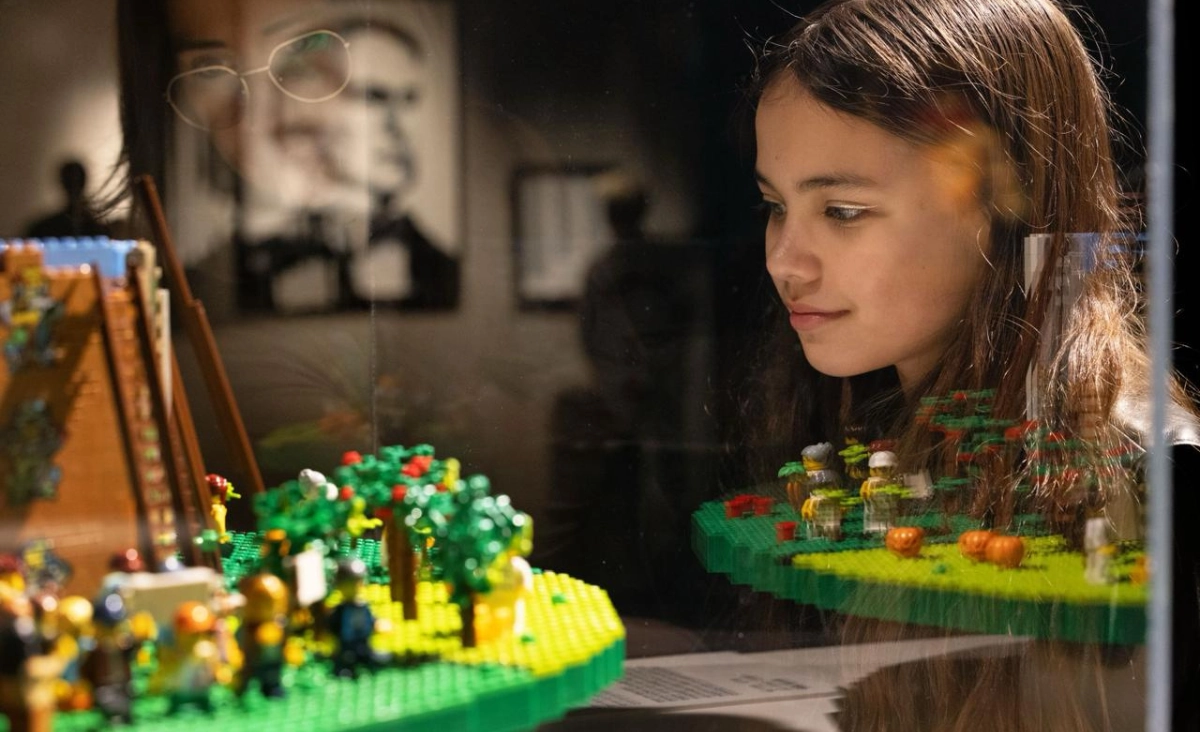 Junges Mädchen betrachtet konzentriert eine detailreiche LEGO-Landschaft mit Bäumen und Figuren in einer Ausstellung.