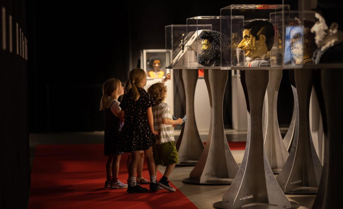 Vier Kinder betrachten in einer Ausstellung mehrere detailreiche LEGO-Kopfmodelle in transparenten Vitrinen auf weißen Sockeln.