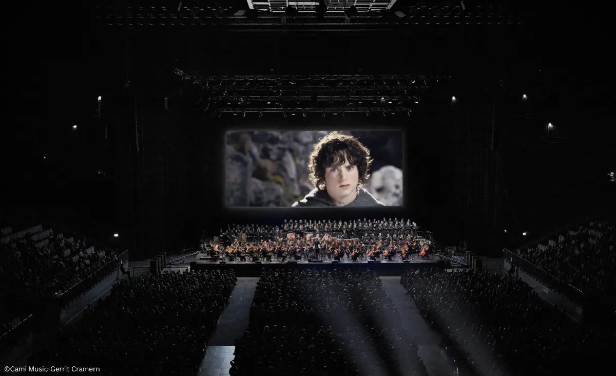 Großes Konzertpublikum vor einer Bühne mit Orchester und einer Leinwand, die eine Nahaufnahme von Frodo aus Der Herr der Ringe zeigt