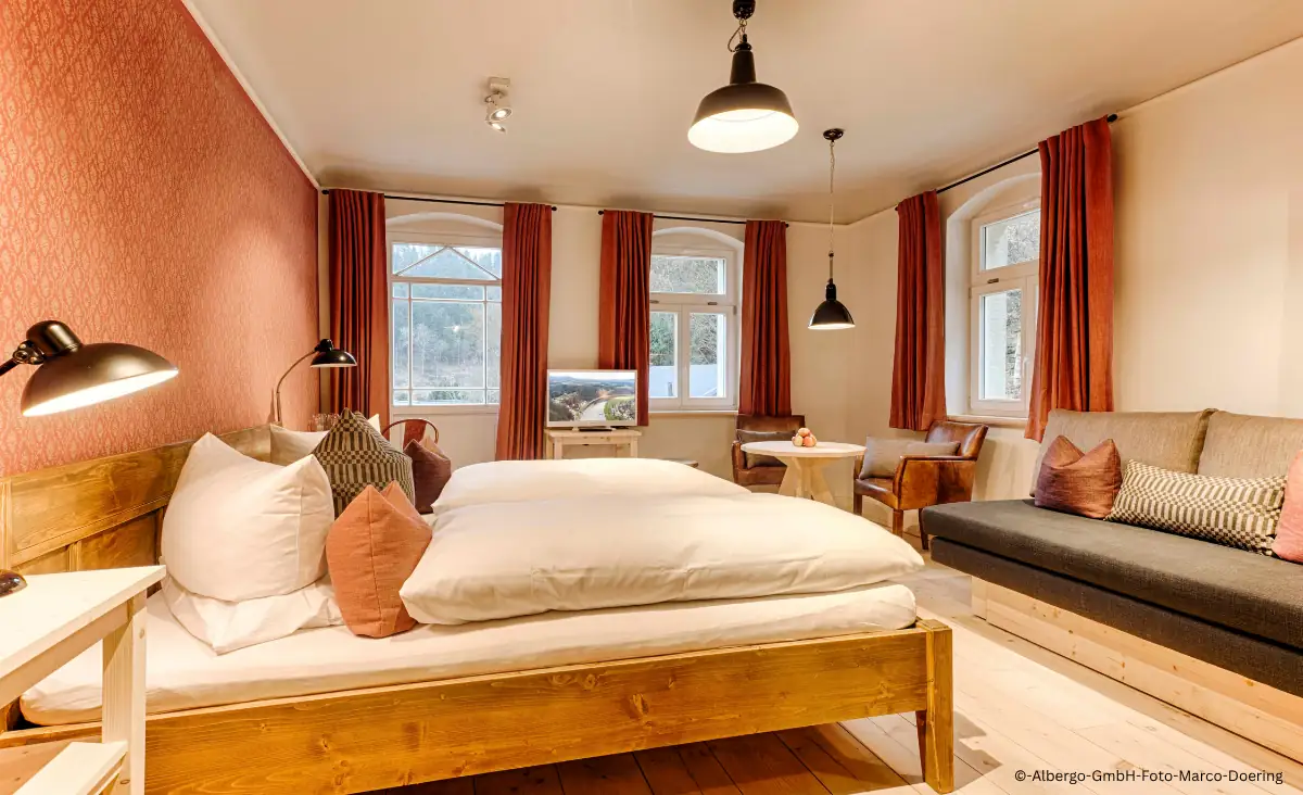 Hotelzimmer mit Holzbett, Sofa, Sesseln, großen Fenstern und orangen Vorhängen im Winterdorf Schmilka Hotel Zur Mühle